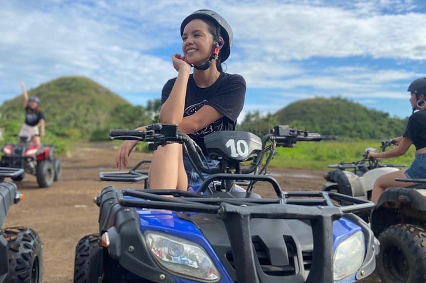 A person on a blue ATV with the number 10, another person on a red ATV in the background, and a hilly landscape. 2