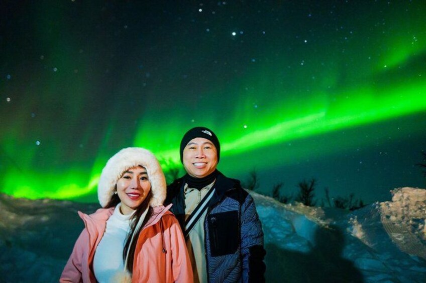 Twee personen staan in een besneeuwd landschap, met het noorderlicht op de achtergrond. 2