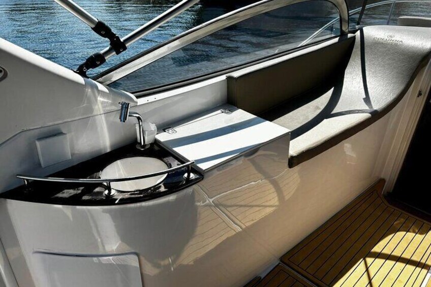 Interior de un barco con un lavamanos de metal, un asiento acolchado y una ventana con vistas al agua. 2