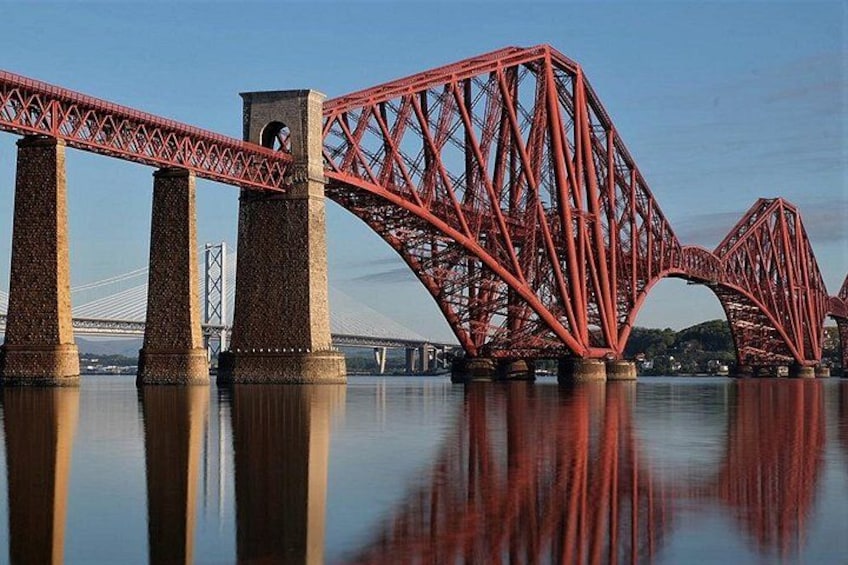 A red steel bridge with a complex truss design spanning over water, with a clear reflection in the water below. 2