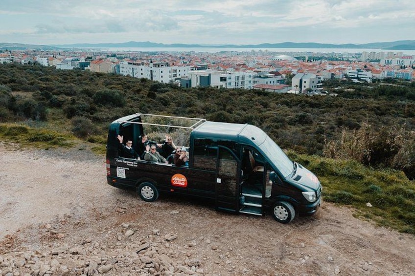 En svart skåpbil står parkerad på en grusväg, full med passagerare. Omgivningen består av ett kuperat landskap och en stad syns i bakgrunden. 4