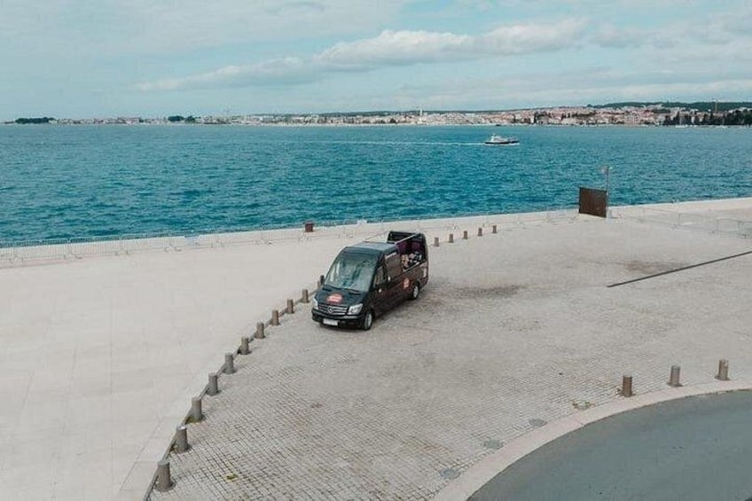En svart skåpbil står parkerad på en asfalterad strandpromenad, med en vattenyta och avlägsna byggnader i bakgrunden. 3
