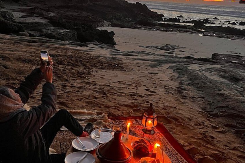 A person sitting on a beach at sunset, holding a smartphone, with a table set for a meal and candles. 5