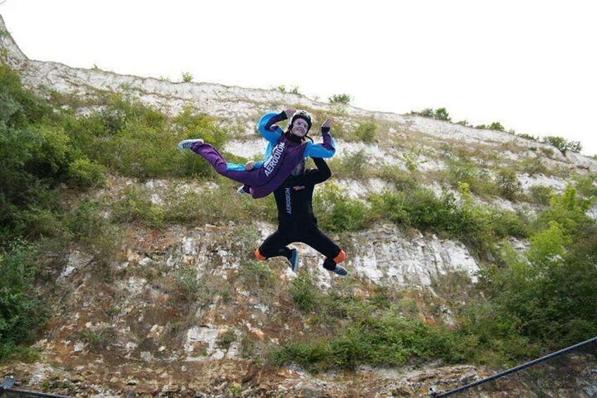 A person in a blue and purple jumpsuit mid-air, possibly jumping or falling, with a rocky hillside in the background. 3