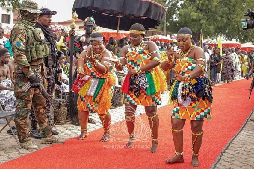 A group of people in colorful traditional attire on a red carpet, with military personnel in the background. 4