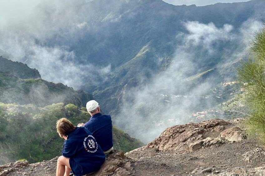 Two people sitting on a rocky outcrop overlooking a mountainous landscape with clouds. 4