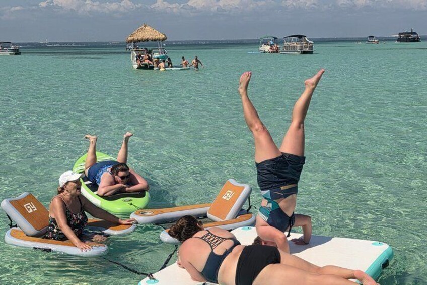 People relaxing on floating loungers in clear turquoise water, with boats and a thatched structure in the background. 3