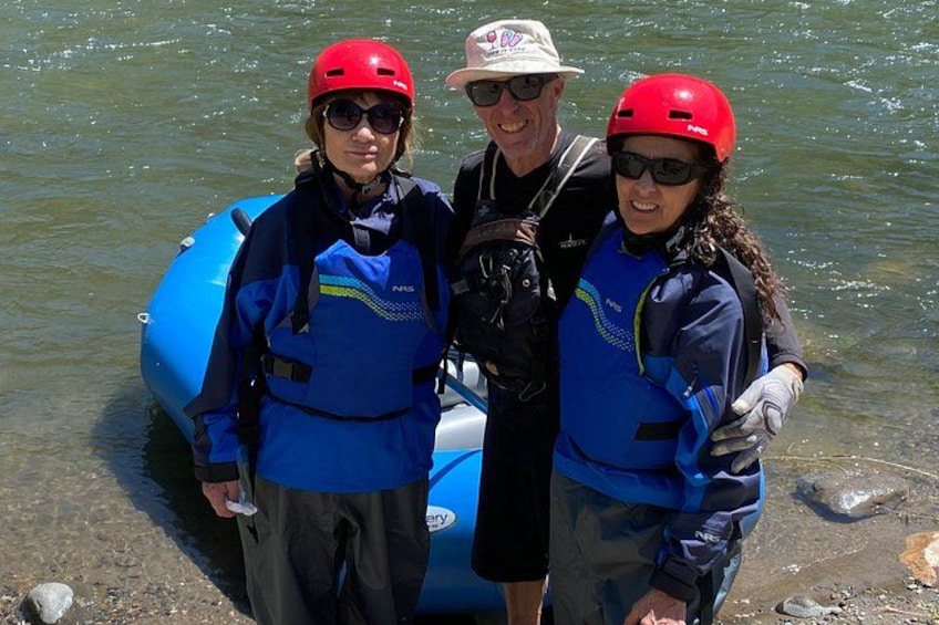 Three people wearing blue life jackets and red helmets standing next to a blue inflatable raft on a riverbank. 4