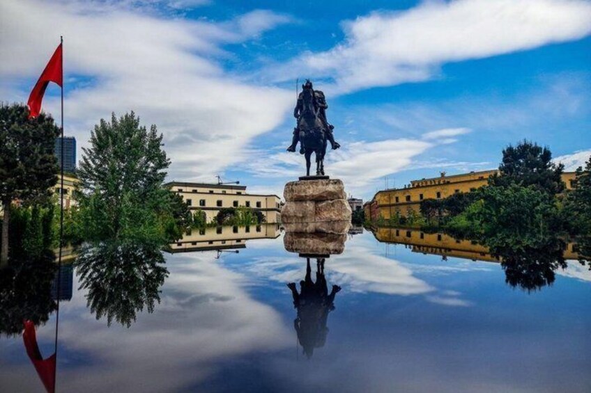 A statue on a pedestal, a red flag, and buildings reflected in a body of water. 4