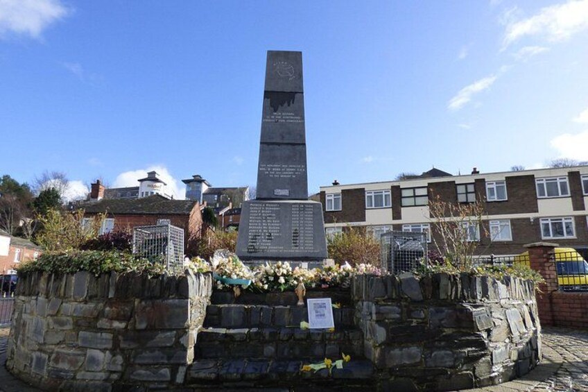 Et højt monument med inskriptioner, omgivet af stenvægge og blomstrende planter. 3