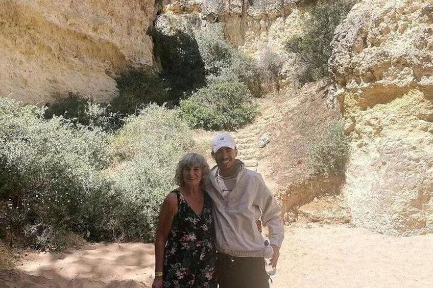 Two people standing on a dirt path with rocky cliffs and sparse vegetation in the background. 3