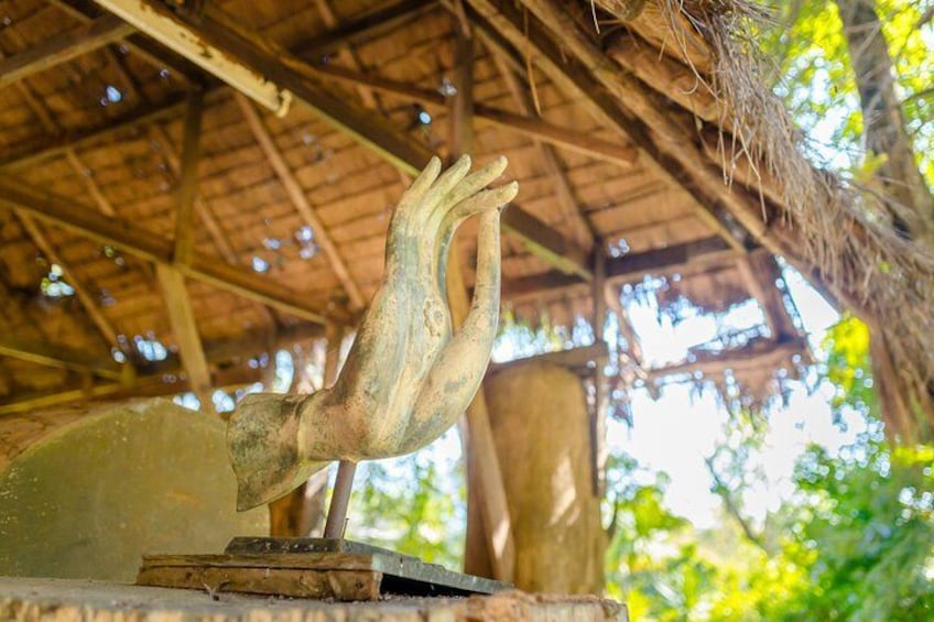 A rustic wooden sculpture of a hand reaching upwards, set against a backdrop of a thatched roof structure and lush greenery. 5