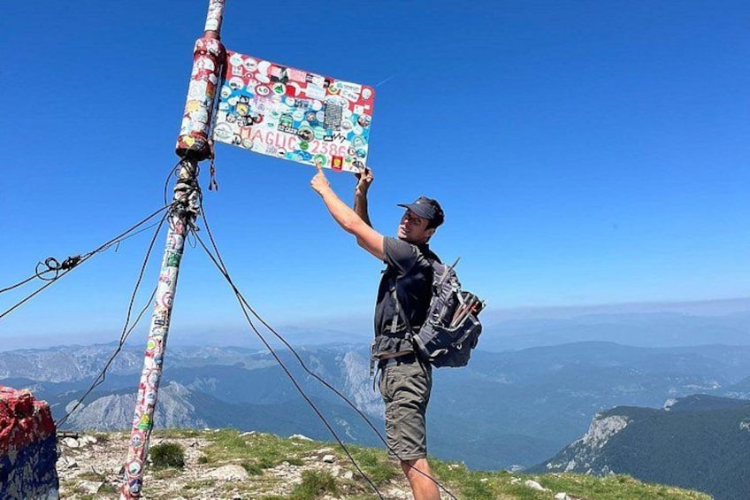 Eine Person steht auf dem Berggipfel mit einer Flagge, einem Rucksack und einem wolkenlosen, blauen Himmel. 4