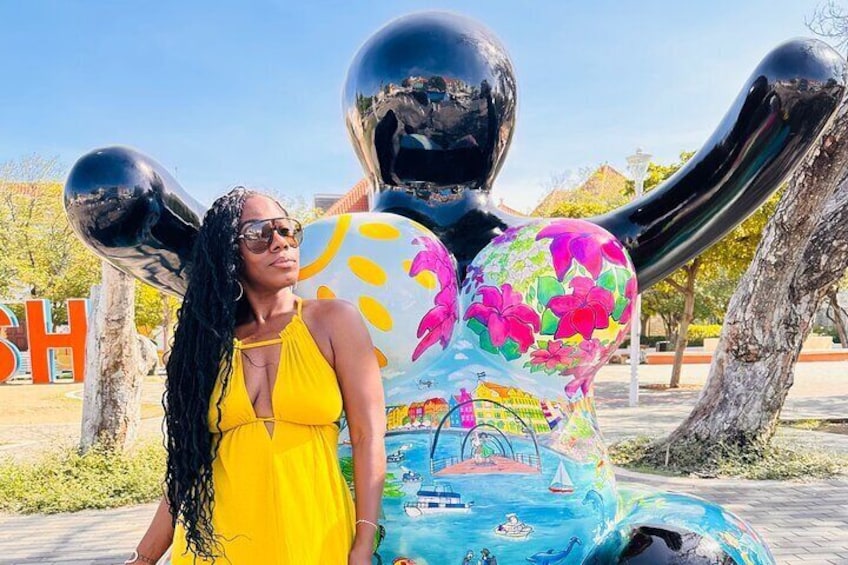 Une personne vêtue d’une robe jaune se tient à côté d’une sculpture colorée représentant un voilier et un arc-en-ciel. 5