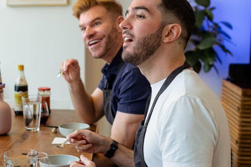 Two people sitting at a table with a bottle of sauce, a bowl, and glasses. 3