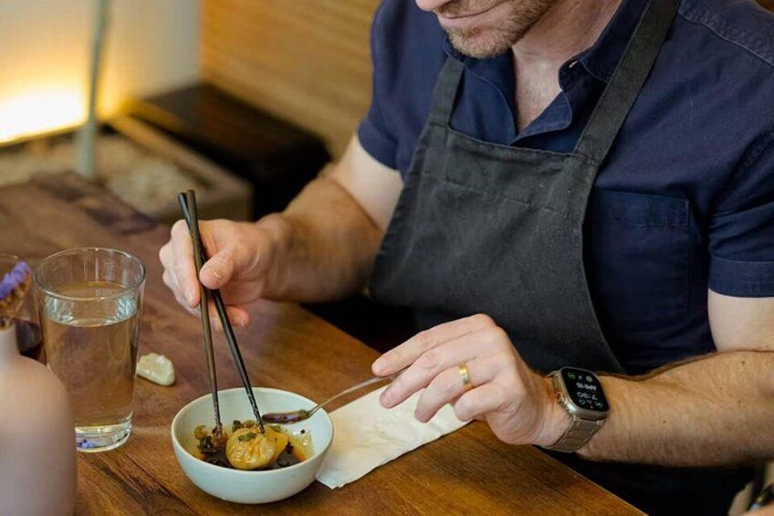 A person wearing a dark blue shirt and a dark apron, holding chopsticks over a bowl of food. 2