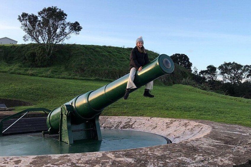 A person sitting on a large green cannon in a grassy area with a hill and trees in the background. 5