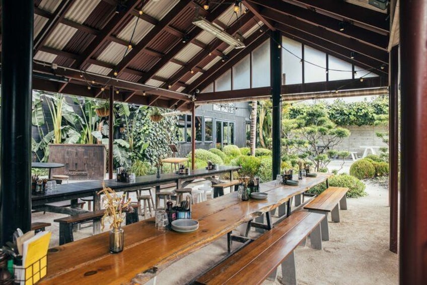 An outdoor cafe with wooden tables and chairs, surrounded by greenery and a covered roof. 4