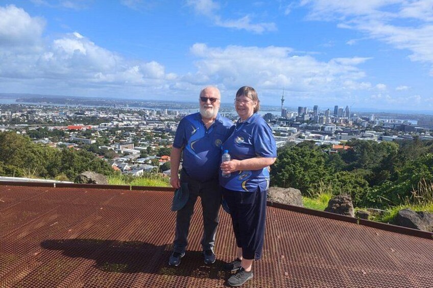 Two people standing on a metal platform overlooking a cityscape with a prominent tower. 3