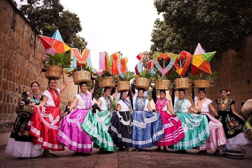 Un groupe de femmes, vêtues de robes traditionnelles aux couleurs vives, porte des paniers sur la tête et se tient devant un mur de pierre orné de drapeaux. 5