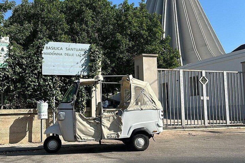 Ein weißes dreirädriges Fahrzeug vor einem von einem Tor geschützten Bereich, auf dem ein Schild mit der Aufschrift 'BASILICA SANTUARIO MADONNA DELLA LACRIME' steht. 4