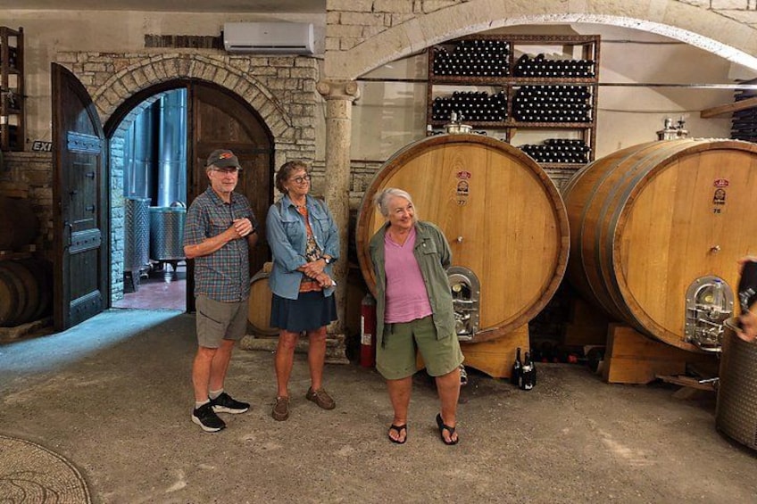Three people standing in a cellar with large wooden barrels and a stone archway. 4