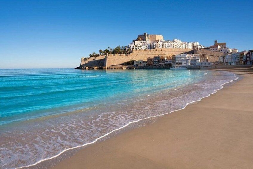 Una playa con agua turquesa cristalina, una orilla arenosa y un pueblo histórico con edificios blancos en una colina. 2