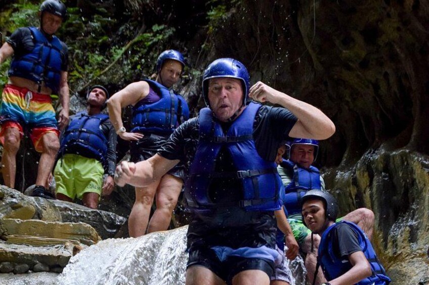 Een groep mensen in blauwe redingswesten en helmen maakt zich klaar om een rotsachtige waterval te abseilen. 4