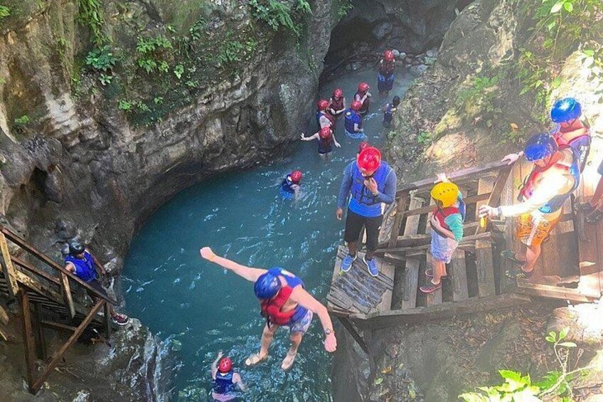 Een groep mensen met blauwe helmen en reddingsvesten die zich klaarmaakt om van een houten brug in het water te springen. 2