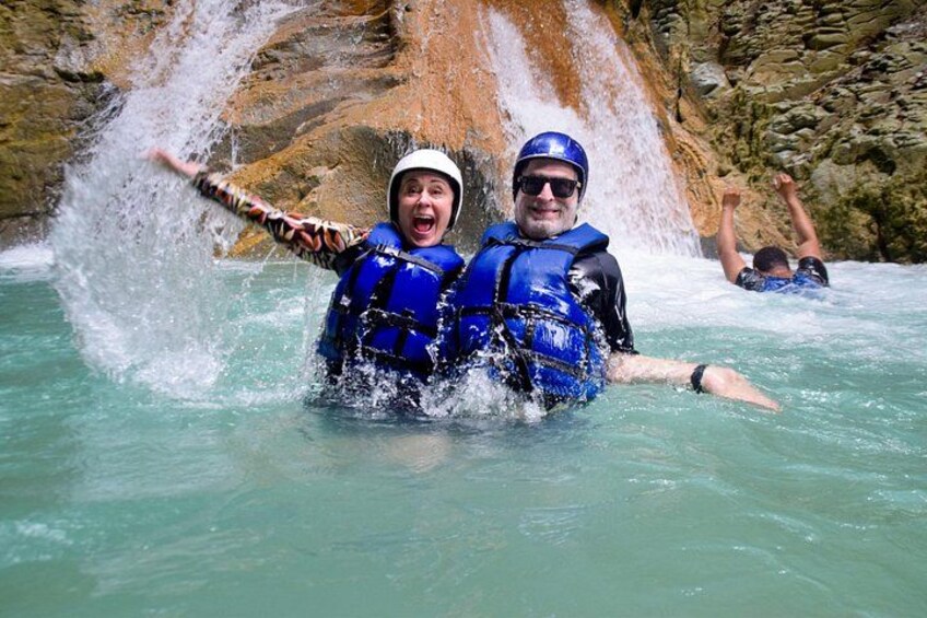 Twee mensen in blauwe reddingsvesten en helmen in een rivier, met een waterval op de achtergrond. 3