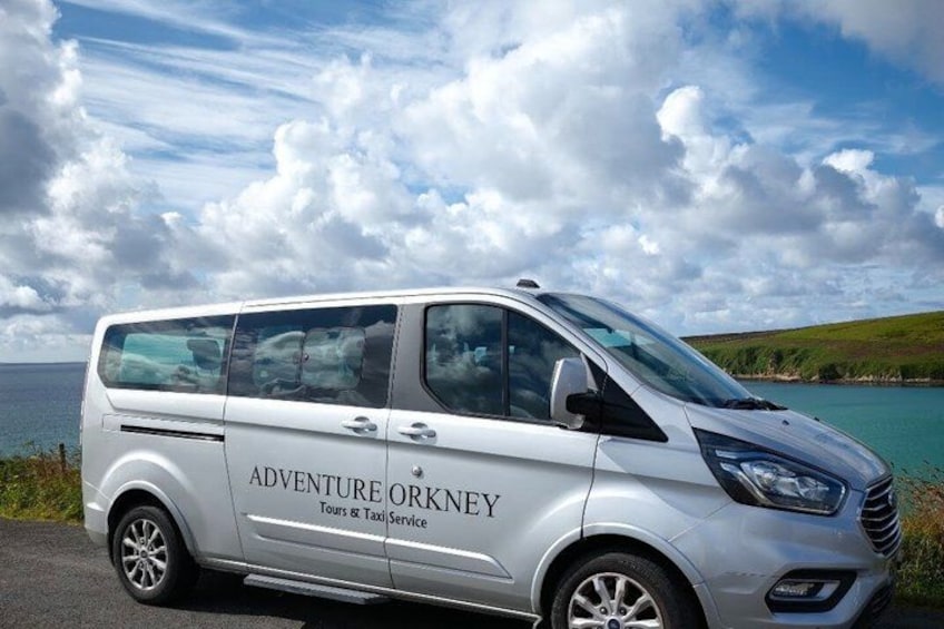 A silver van with 'Adventure Orkney Tours & Taxi Service' written on its side, parked by a lake with a cloudy sky. 3