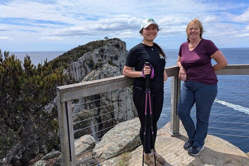 Two people standing on a wooden platform with a railing, overlooking a rocky coastline and the sea. 4