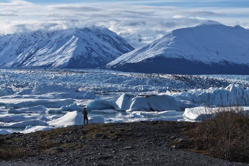 A person standing on a rocky outcrop overlooking a glacier with mountains in the background. 1