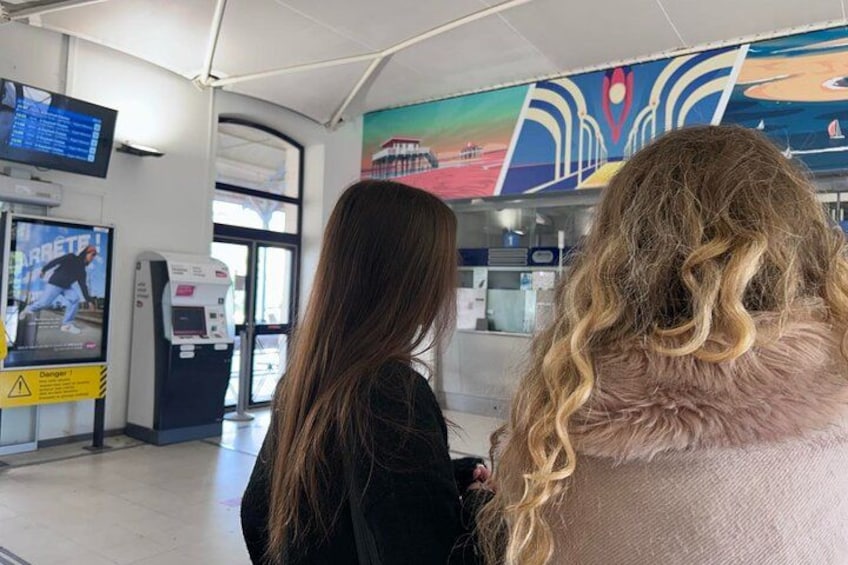 En undergrundsstation med et digitalt informationsskilt, billetautomater og et farverigt muralmaleri på væggen. 3