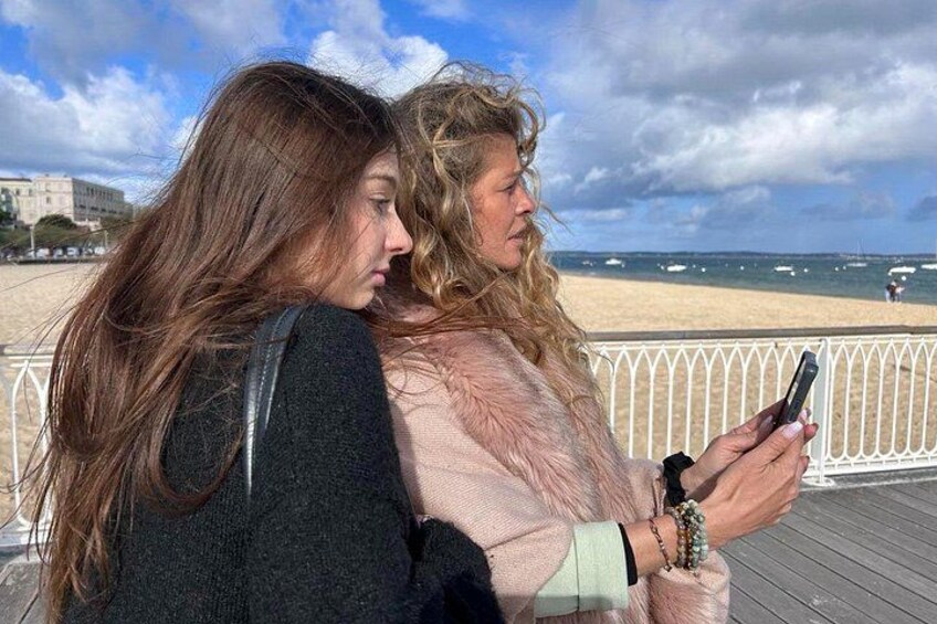 Deux personnes se trouvent sur une promenade en bois au bord de la plage, avec l’océan et des bateaux en arrière-plan. 2