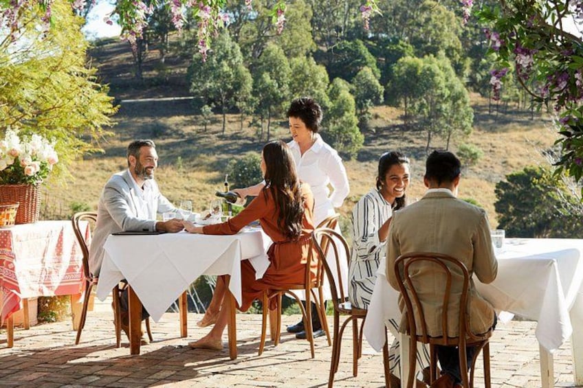 Un groupe de personnes est assis à une table à l’extérieur, recouverte d’une nappe blanche, et entourée de chaises. L’ensemble se trouve dans un jardin, avec des arbres et des collines en arrière-plan. 2