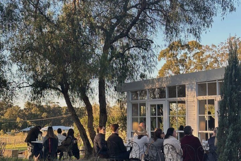 Un gruppo di persone riunite attorno a un edificio con ampie finestre, immerso in un paesaggio naturale con alberi e cielo sereno. 1