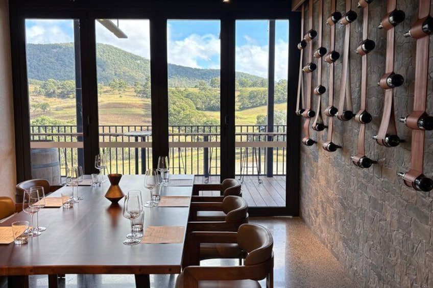 Un tavolo da pranzo con bicchieri da vino, una sedia di legno e vista su un vigneto attraverso ampie finestre. 2