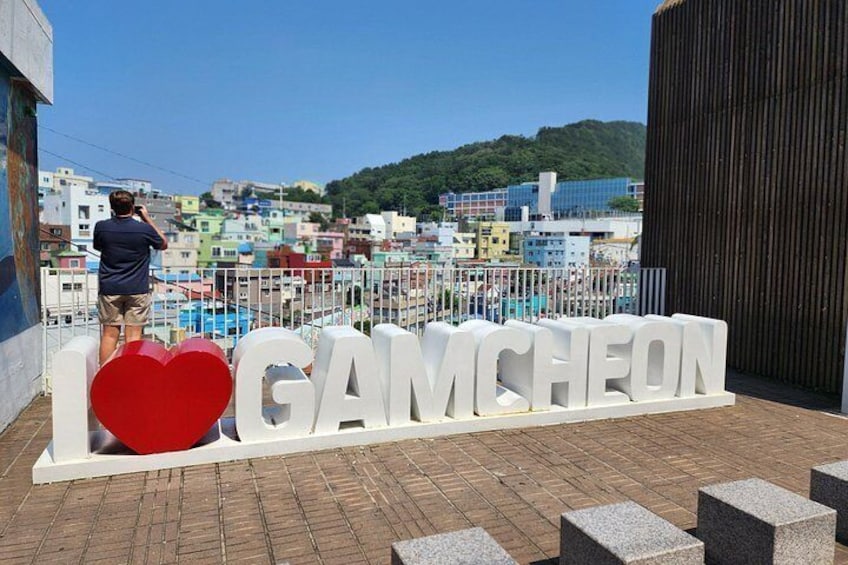A person taking a photo, a large white sign with 'I ♥ GAMCHEON' and a red heart, and a cityscape in the background. 4
