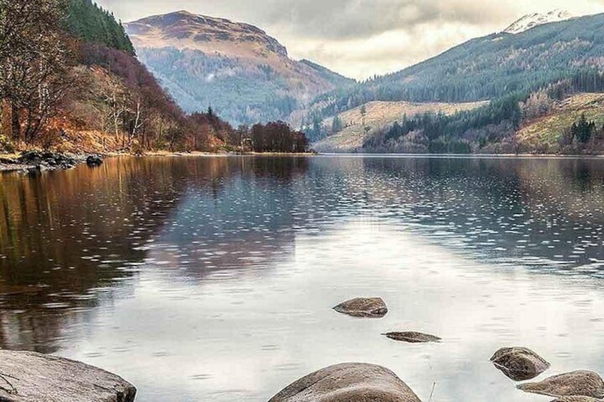 A calm lake surrounded by mountains, with rocks in the foreground and trees along the shore. 2