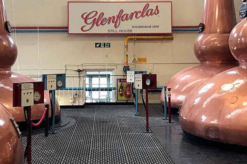 A distillery interior with large copper stills, a sign reading 'Glenfarclas Still House,' and a check-in counter. 2