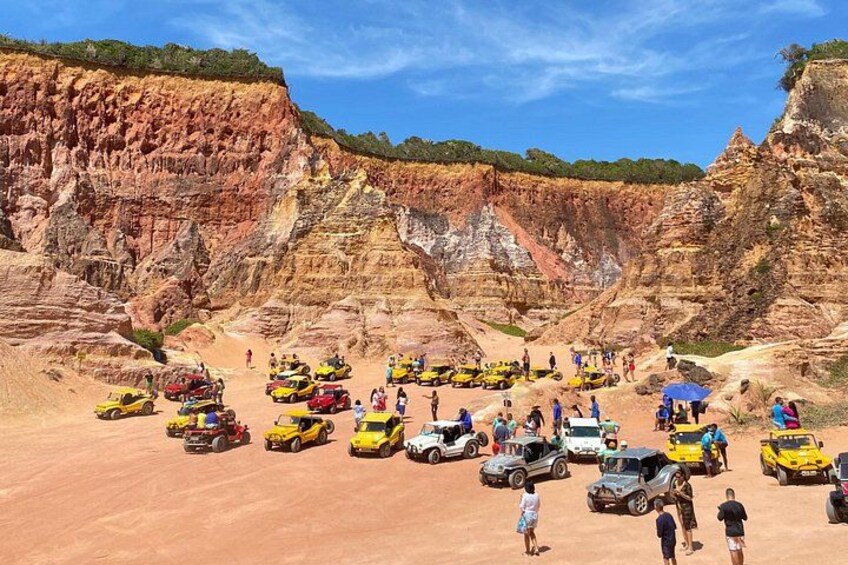 A group of people and vehicles on a desert-like terrain with layered rock formations in the background. 4