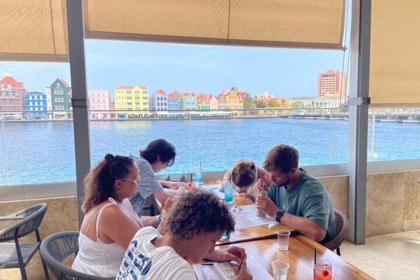 A group of people dining at a table with a view of a waterfront and colorful buildings in the background. 1