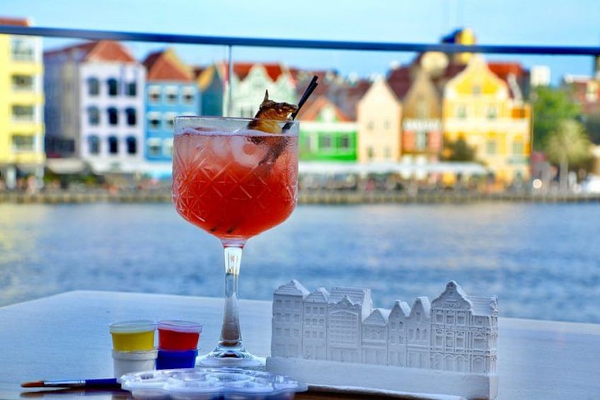 Un vaso de cóctel con una bebida roja, una pequeña escultura y vasos coloridos sobre una mesa junto a un muelle, con edificios coloridos al fondo. 5