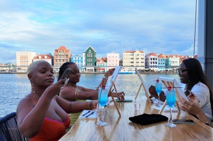 Three people sitting at a table with drinks, overlooking a waterfront with colorful buildings. 1