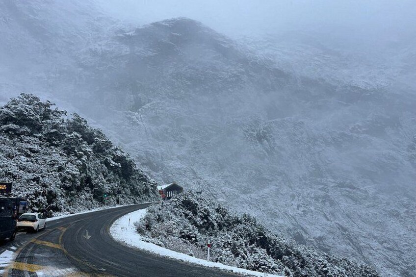 A snow-covered mountain road with a curve, a white car, and a small building. 2