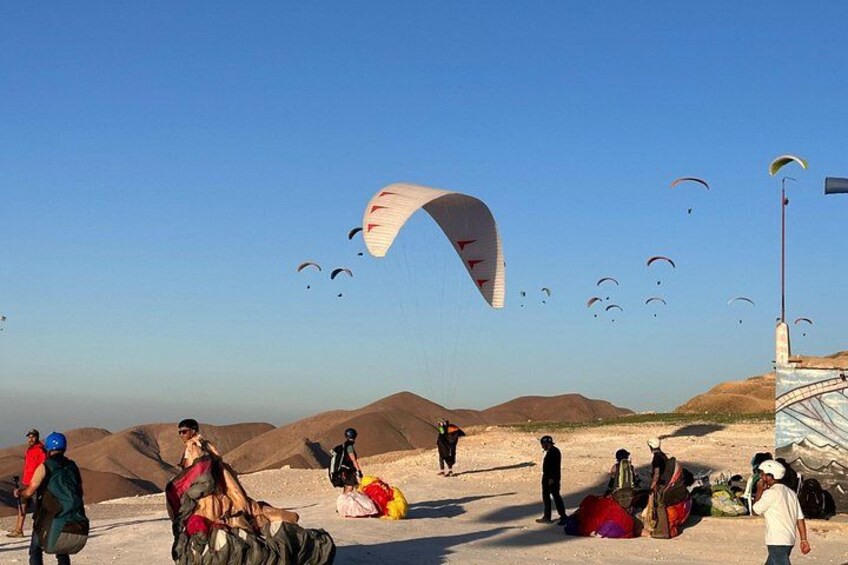 A group of people preparing for paragliding with several parachutes visible in the sky. 1