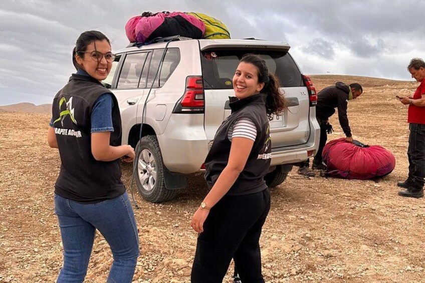 Two people standing next to a vehicle with a large bag on top, and another person setting up a red bag on the ground. 2