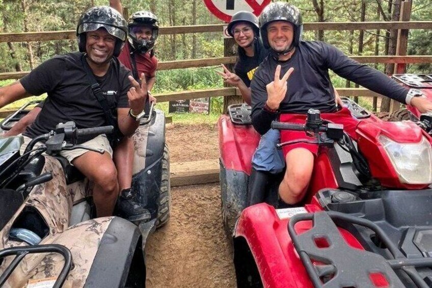 Quatre personnes à bord de quads, une sur un quad rouge et trois sur un quad beige, avec une clôture en bois et des arbres en arrière-plan. 1