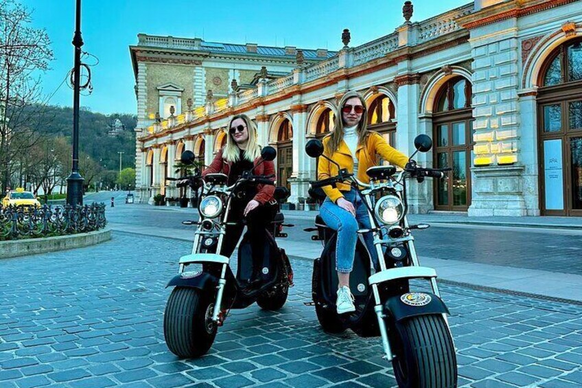 Two people riding electric scooters on a cobblestone street in front of a historic building. 4